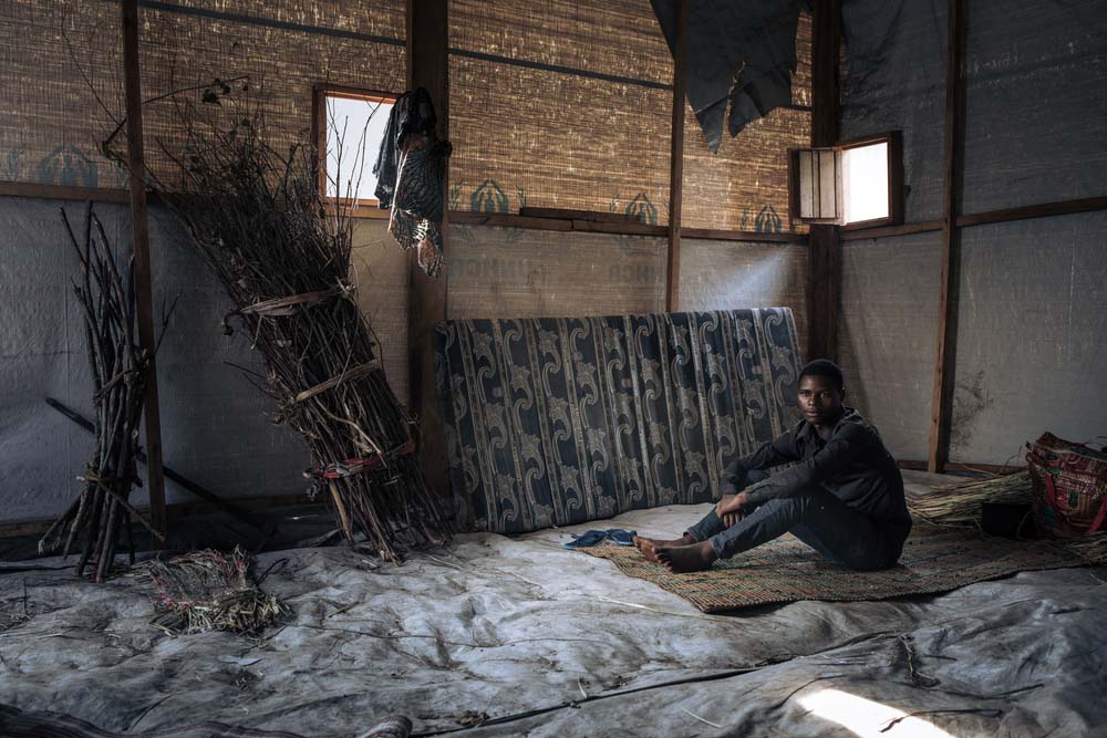 bed, a 20-year-old, single, no children, displaced man, stands with his only belongings in the community shelter where he lives in the informal IDP site of Rugabo Stadium in Rutshuru Centre, in North Kivu province, eastern Democratic Republic of Congo.