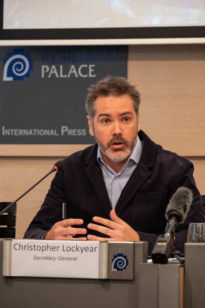 Christopher Lockyear, Secretary General of Doctors Without Borders (MSF), speaks during a press conference at the Residence Palace International Press Centre in Brussels. He is seated at a desk with microphones in front of him, gesturing with his hands while addressing the media.