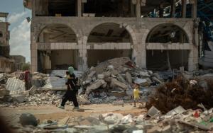 A mother carries a young child in her arms while another young child walks behind her, passing in front of a demolished building in Gaza.