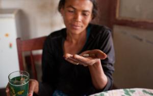 A woman sits at a table, In her hand is medication for HIV/Aids.