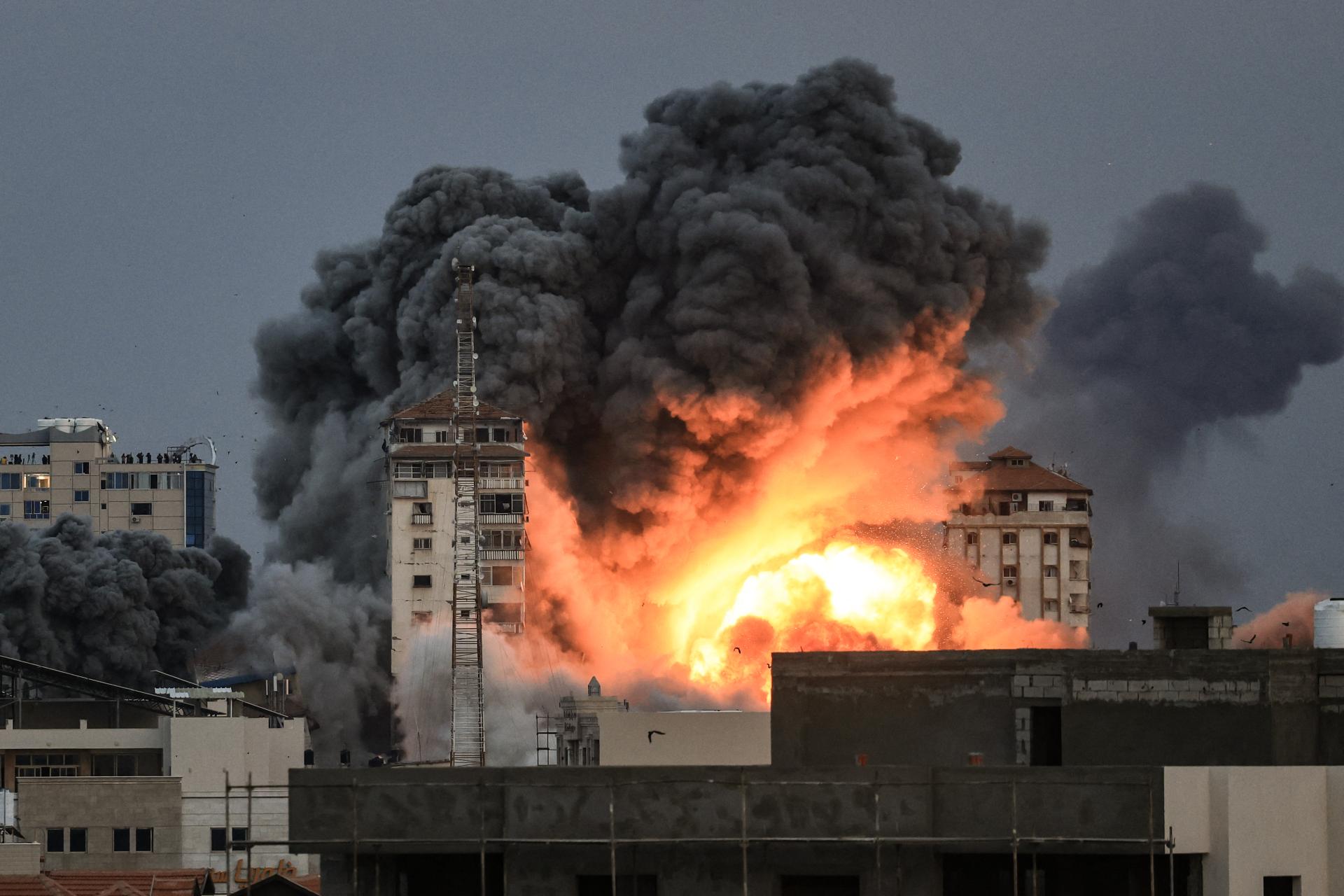 Image showing people standing on a rooftop watch as a ball of fire and smoke rises above a building in Gaza City on October 7, 2023 during an Israeli air strike that hit the Palestine Tower building.