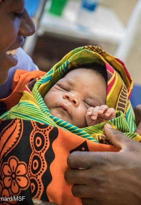 Child health starts early. A newborn is held, wrapped in an orange blanket, while a nurse checks him.
