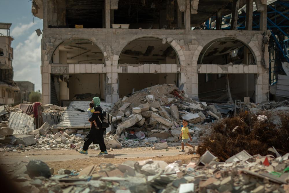 A mother carries a young child in her arms while another young child walks behind her, passing in front of a demolished building in Gaza.