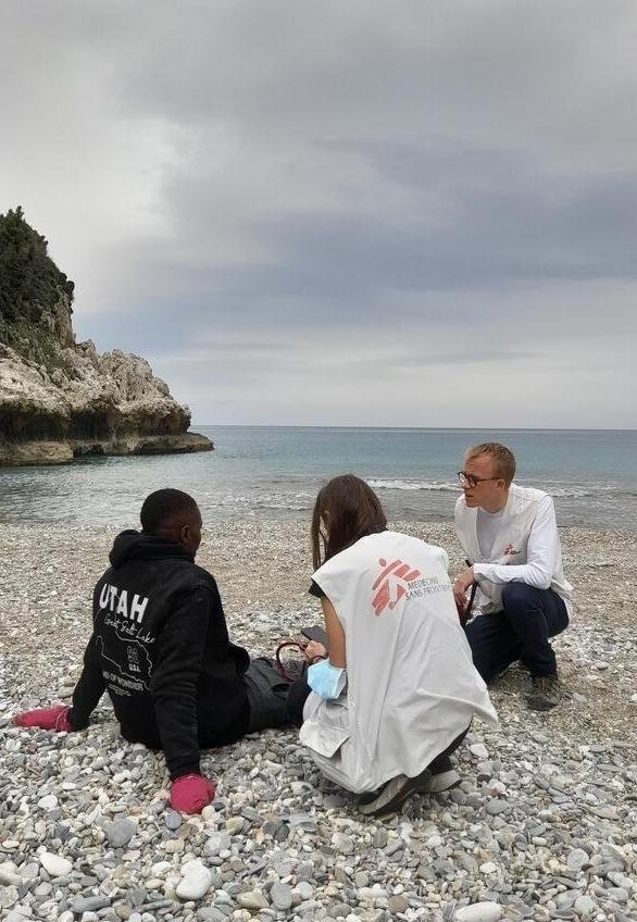 A man is sitting on a beach, catching his breath, while an MSF doctor is examining him and chatting about his mental health. 