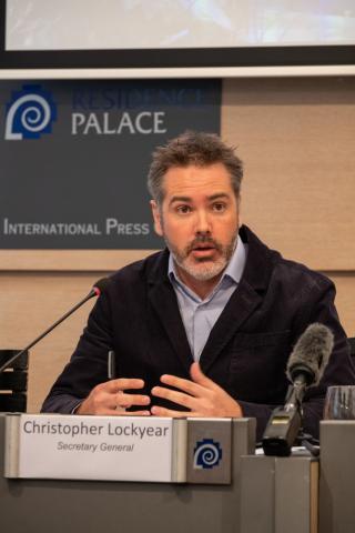 Christopher Lockyear, Secretary General of Doctors Without Borders (MSF), speaks during a press conference at the Residence Palace International Press Centre in Brussels. He is seated at a desk with microphones in front of him, gesturing with his hands while addressing the media.